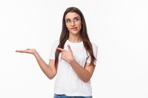 Waist-up portrait of thoughtful, pretty young woman holding arm as if have something lying in it, pointing at product with pondering face, bite lip look up thinking, white background photo