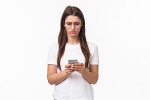 Communication, technology and lifestyle concept. Portrait of troubled and confused young woman reading strange message, frowning as looking mobile phone display, stand white background photo