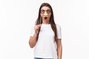 Entertainment, fun and holidays concept. Funny and cute young girl fool around, praying wearing glasses and nose mask, gasping amazed, look surprised standing white background photo