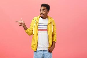 Man did not expect that coming. Portrait of surprised joyful and carefree stylish modern dark-skinned male in yellow jacket with beard and curly hair pointing and gazing left amazed over pink wall photo