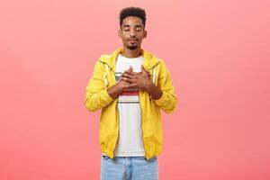 Attractive and stylish dark-skinned guy feeling warm and calm feelings inside holding hands on heart closing eyes and smiling with relieved comfort expression standing over pink wall in trendy outfit photo