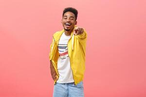 ha-ha loser. Portrait of happy amused energetic african american bearded male in yellow trendy jacket pointing at camera with index finger and laughing out loud humiliating mocking strange weirdo photo