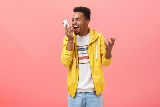 Guy being fed up with dumb customer support shouting at smartphone screen yelling from pissed outrage feelings clenching gist holding cellphone near face over pink background photo