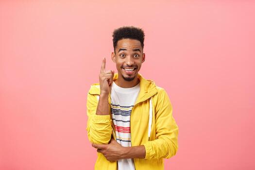 Enthusiastic pleased african american guy adding suggestion raising index finger in eureka gesture and smiling joyfully while discussing interesting invention or theory smiling broadly over pink wall photo