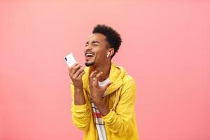 Singing from bottom of heart along favorite music listening song in wireless earbuds with closed eyes and joyful smile gesturing passionately making microphone from smartphone over pink background photo