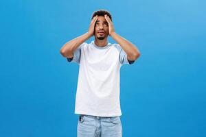 Man feeling reckelss cannot understand why bad things happen him. Distressed panicking and anxious miserable dark-skinned young man holding hands on head looking up in sky being in tough situation photo