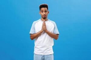 Please help me beg you. Charming cute and handsome african american boyfriend with afro curly haircut and beard holding hands in pray frowning making silly face while asking favor over blue wall photo