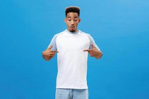 Impressed emotive african american male with beard and afro hairstyle folding lips in wow sound pointing at chest or centre with amazement as if looking at awesome copy space over blue background photo