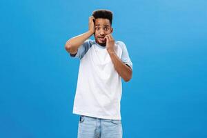 Insecure anxious guy getting scared of consequences. Portrait of frightened panicking handsome young african american male biting fingernails gazing worried at camera posing over blue background photo