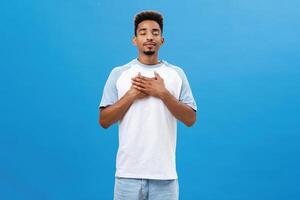 Guy finding peace in poetry breathing fresh air closing eyes with peaceful relaxed expression smiling holding palms on heart relieving from stress posing over blue background photo