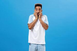 Portrait of frightened insecure dark-skinned guy having fobia biting fingernails raising eyes and trebmling from fear and fright standing scared over blue background in casual summer t-shirt photo