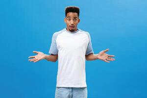 Handsome intense dark-skinned guy feeling concerned and questioned having no clue what happened being worried spreading palms aside with confused expression standing uncertain over blue background photo