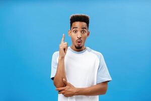 Artistic and creative dark-skinned boyfriend in trendy t-shirt folding lips in wow, raising index finger in eureka gesture making up great idea adding suggestion how solve situation over blue wall photo