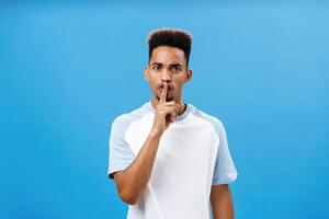Waist-up shot of bothered and pissed good-looking african man with afro hairstyle frowning from irritation and annoyance demanding shut up keep secret and shushing at camera over blue wall photo