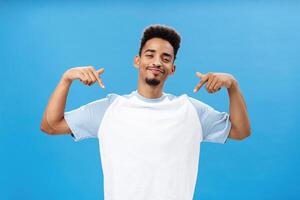 Yeah look how cool I am. Proud arrogant and stylish dark-skinned man with beard squinting and smirking with delight pointing at himself talking about personal achievements over blue background photo