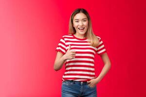 Nice idea my approval. Pleased satisfied good-looking blond asian girl show thumb up and smile, give positive reply, agree wish good luck, judge excellent work, stand red background photo