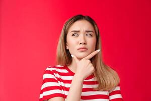 Close-up suspicious thoughtful girlfriend pondering, touch chin squinting doubtful and frowning, look up while thinking, take decision, stand red background perplexed, hesitating making choice photo