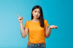 Troubled cute upset asian girl have problem asking your help, pointing upper left corner shrugging unsure, look frustrated and sad, questioned answer, cannot deal problem, stand blue background photo