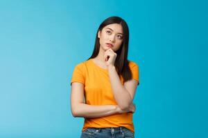 Thoughtful dreamy smart asian girl thinking, making plan inside head, touch chin, look up focused, contemplating interesting scene, pondering, making choice, taking decision, stand blue background photo