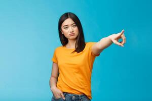 Go away from here. Serious-looking arrogant displeased bossy asian girl showing way-out, pointing sideways index finger look camera demand leave, stand blue background ignorant photo