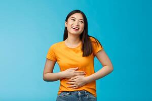 Happy staffed cute asian girl feelings satisfaction after eating favorite food, smiling broadly rubbing tummy, touch stomach from delight and joy, like eat delicious meals, stand blue background photo