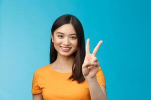 Cheerful friendly asian woman in yellow t-shirt smiling broadly, show number two, twice, win second place, smiling joyfully, make take-away order, book sits, stand blue background photo