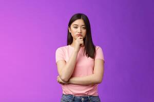 Judgemental serious-looking smart asian girl give expert look, touch chin thoughtful, squinting suspicious uncertain, make decision, choosing product, thinking, pondering plan, purple background photo