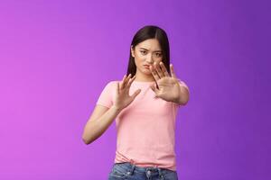 Displeased intense reluctant asian girl blocking, trying protect face, raise hands stop, prohibition gesture, frowning sulking, refusing bothering offer, stand purple background, rejecting photo