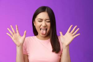 Close-up silly moody asian girl complaining act immature and bothered, raise hands unsatisfied show tongue aversion disgust, close eyes, react terrible disgusting thing, stand purple background photo