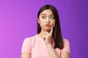 Close-up thoughtful cute asian girl dreamy standing purple background, touch cheek, turn left, thinking, doubting how act, wondering information, contemplating view from window photo
