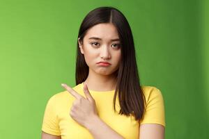Grumpy upset cute asian girl complaining tired, drained during university exams, pointing unhappy, losing competition, pointing left, sulking displeased, stand green background reluctant photo