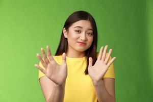 Modest cute insecure timid asian girl waving hands no gesture, step back, smirk, cringe aversion and dislike, refusing suspiciosu offer, rejecting proposal, stand green background photo