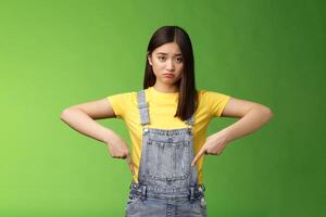 Sad asian girl in dispair feel upset, frowning sulking pessimistic, pointing down look camera pulling sorrow uneasy face, express sadness and frustration, stand green background photo