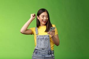 Triumphing cute joyful asian girl hold smartphone, fist pump celebrating good news, read good news message, look telephone screen smiling happily, achieve success got persmission party photo