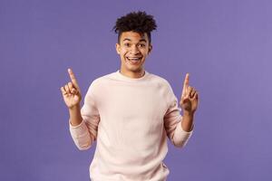 Portrait of charismatic lively young man heard about good deal, asking question intrigued, pointing fingers up, smiling upbeat at camera, look with interest, recommend product, purple background photo
