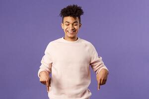 Portrait of cute enthusiastic young man found something really cool, pointing fingers down, happy smiling and peeking at bottom advertisement with pleased grin, purple background photo