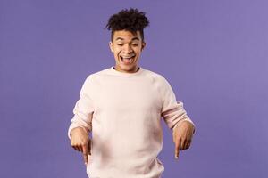 Joyful good-looking hipster guy with dreads rejoicing over good news, best deal ever in online store, favorite game finally in stock, pointing fingers down and rejoicing, purple background photo