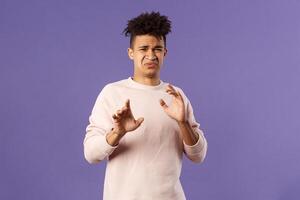 Phew get it away from me. Portrait of disgusted young man smelling something awful, step away and blocking it with raised arms, refuse grimacing with aversion and reluctance, purple background photo