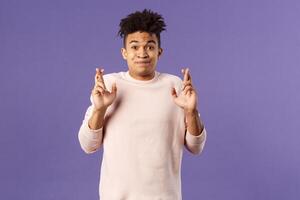 Portrait of young hopeful handsome man with dreads ancitipating something good happen, cross fingers good luck, smiling with joy and optimism, having faith, praying over purple background photo