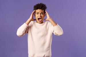 Portrait of scared and anxious young hispanic handsome man in panic, shaking hands nead head, screaming and gasping afraid, being in trouble, standing alarmed over purple background photo