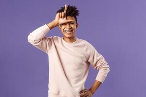 Portrait of charismatic happy, boastful young hispanic guy mocking friend for losing to him, showing loser gesture over forehead and laughing over defeated person, stand purple background photo