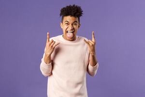 Portrait of enthusiastic, excited funny young man having fun, enjoying awesome rock concert, show heavy metal sign, stick tongue and rejoicing hear favorite band on stage, purple background photo