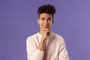 Close-up portrait of indecisive, puzzled young man facing difficult choice, look up thoughtful, touch chin and grimacing while thinking, making-up idea or decision, purple background photo