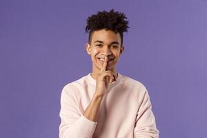 Close-up portrait of attractive smiling young man hiding secret, asking keep silent or quiet to do surprise, show shush gesture, place index finger to mouth, standing purple background photo