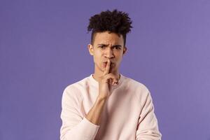 Show some respect. Portrait of angry and annoyed, displeased young hispanic man telling to keep quiet, shushing at someone being too loud, hold index finger over mouth frowning, scolding person photo
