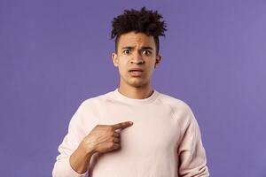Close-up portrait of embarrassed and worried young man being named or called to principal office, pointing at himself and asking why with disbelief and anxiety, standing purple background photo