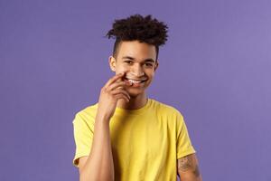 Close-up portrait of lovely young hispanic gay man with dreads, tattoos, touching lip sensually and flirty smiling, checking out someone really cute and handsome, standing purple background photo