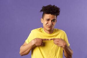 Close-up portrait of shy and modest young silly hispanic man trying say something but being too insecure, grimacing and frowning, look timid, two fingers touching pose, purple background photo