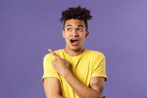 Portrait of impressed hispanic guy seeing something interesting, pointing finger and looking upper left corner, discuss strange thing upwards, standing purple background curious photo