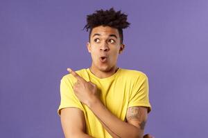 Waist-up portrait of amazed, impressed hispanic male with dreads, seeing something incredible and cool, pointing finger looking upper left corner, breathtaking awesome thing, purple background photo